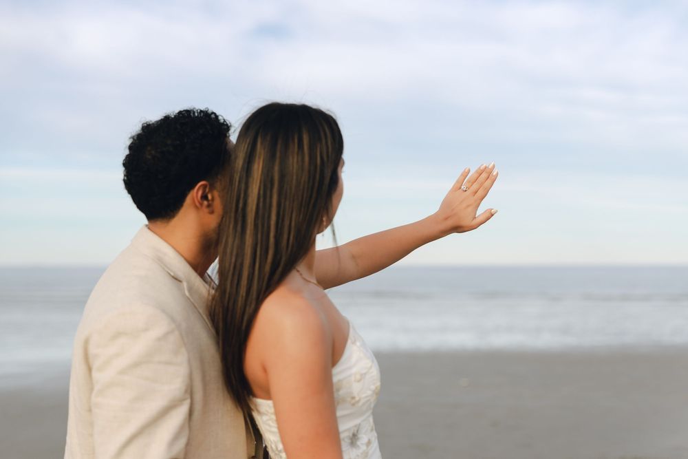 Couple in wedding attire standing on a beach, woman pointing toward the ocean horizon.