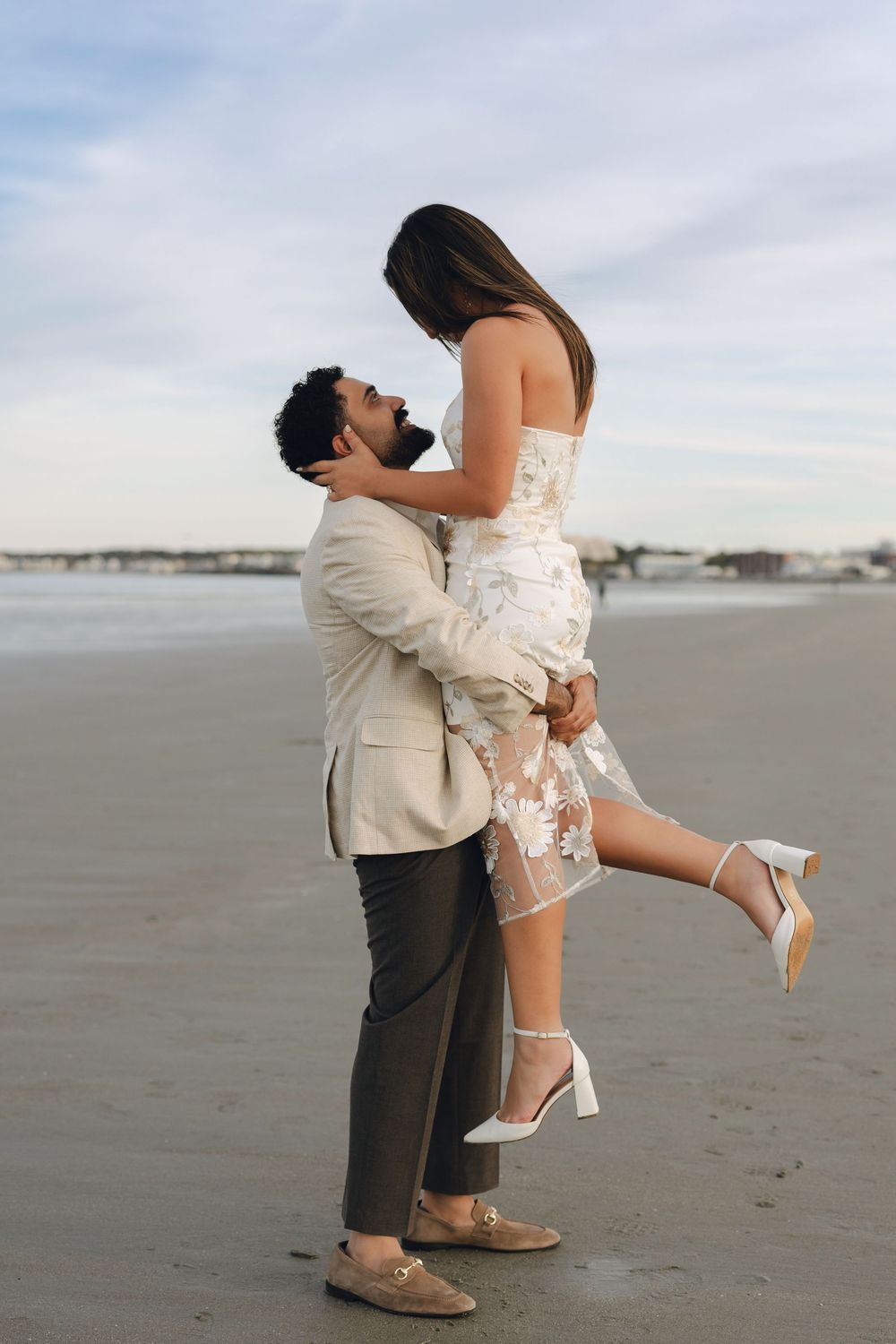 Couple sharing a romantic moment on the beach, man lifting woman in floral dress and heels at sunset.