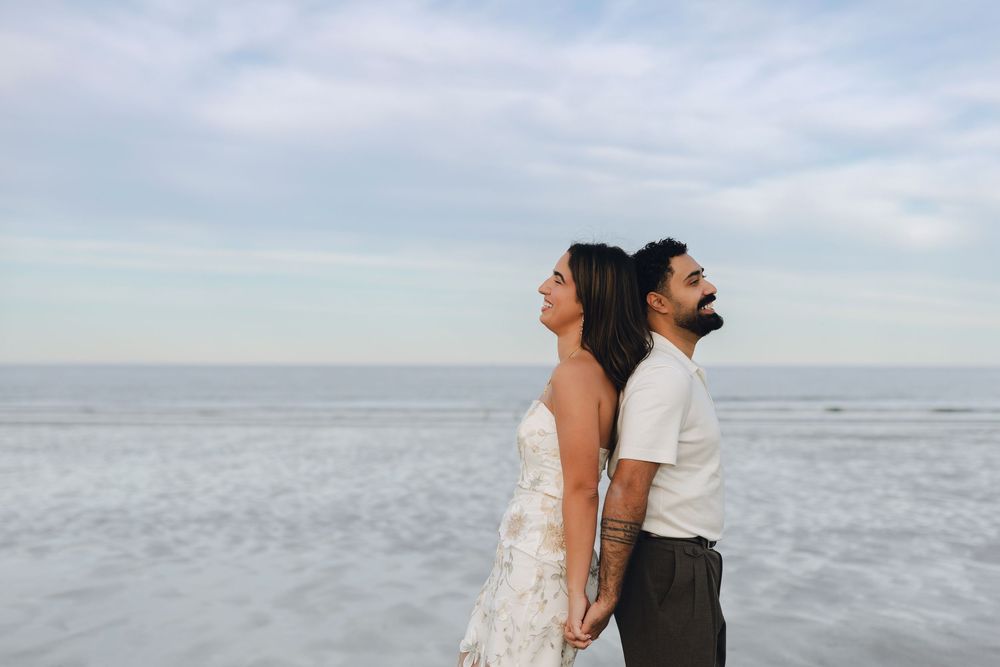 Couple in wedding attire standing back-to-back on a beach, holding hands with ocean waves in the background.