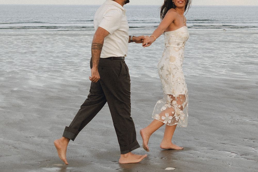 Couple walking barefoot on beach, woman in floral dress holding hands with man in white shirt and dark pants.