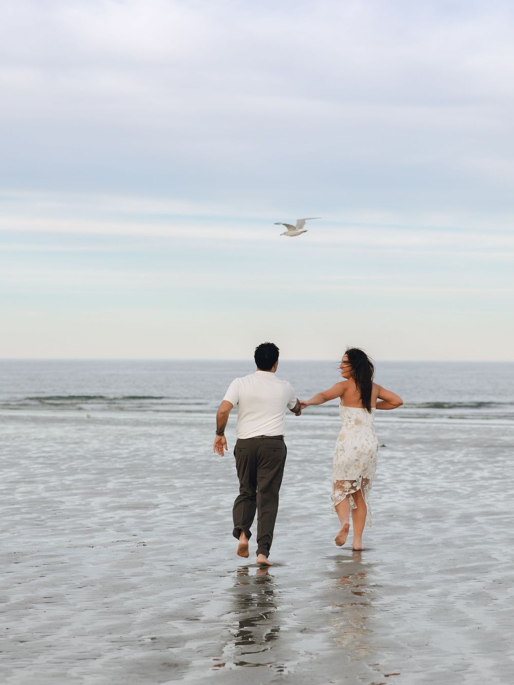A couple runs hand-in-hand along a wet sandy beach toward the ocean with a seagull flying overhead.