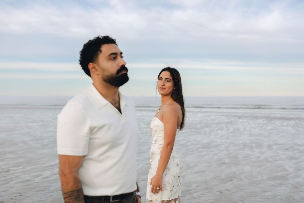 Couple standing on a beach, man in white shirt with tattoos, woman in white dress, ocean in background.