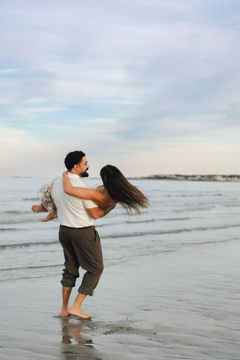 A couple playfully spinning on a sandy beach shore with ocean waves and cloudy sky in the background.