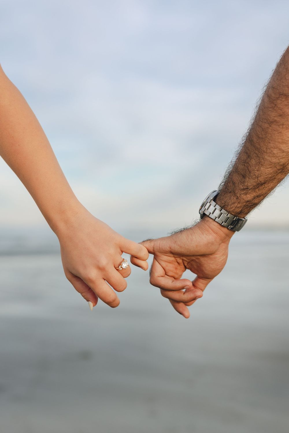 Couple holding pinkies on a beach, woman wearing a diamond engagement ring and man wearing a silver watch.
