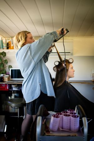Hairdresser styling long brown hair with curlers in a home salon setting.