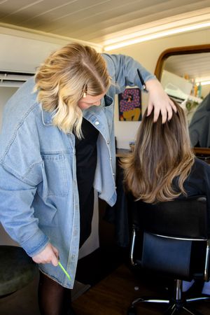 A hairstylist in a denim jacket carefully examines a client's long brown hair in a cozy salon studio.