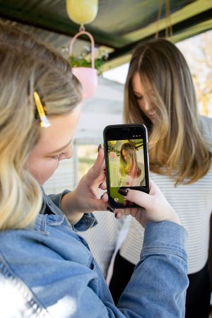 Two young women taking a photo on a smartphone outdoors, with the phone screen showing a preview of the shot.