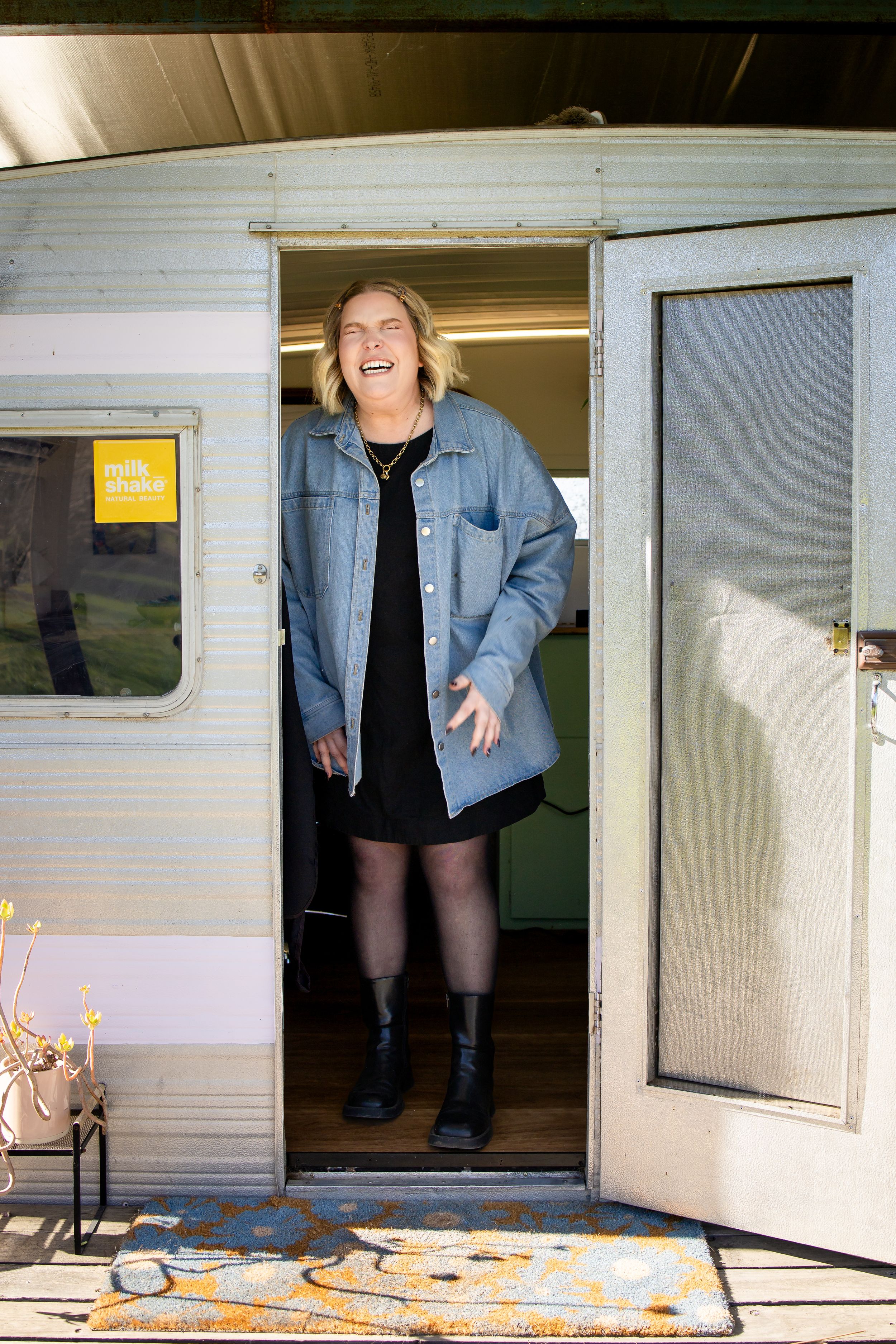 Woman laughing in doorway of vintage caravan wearing denim jacket and black dress with ankle boots.