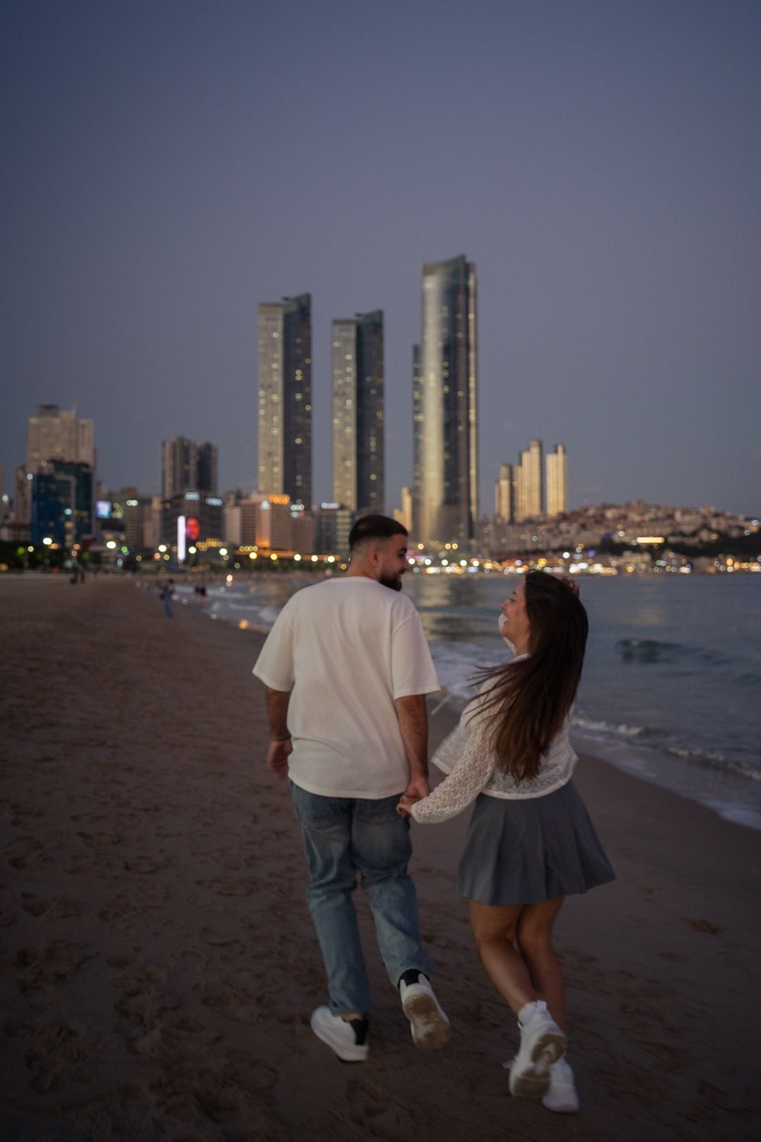 Couple holding hands walking on a sandy beach at dusk with illuminated city skyscrapers in the background.