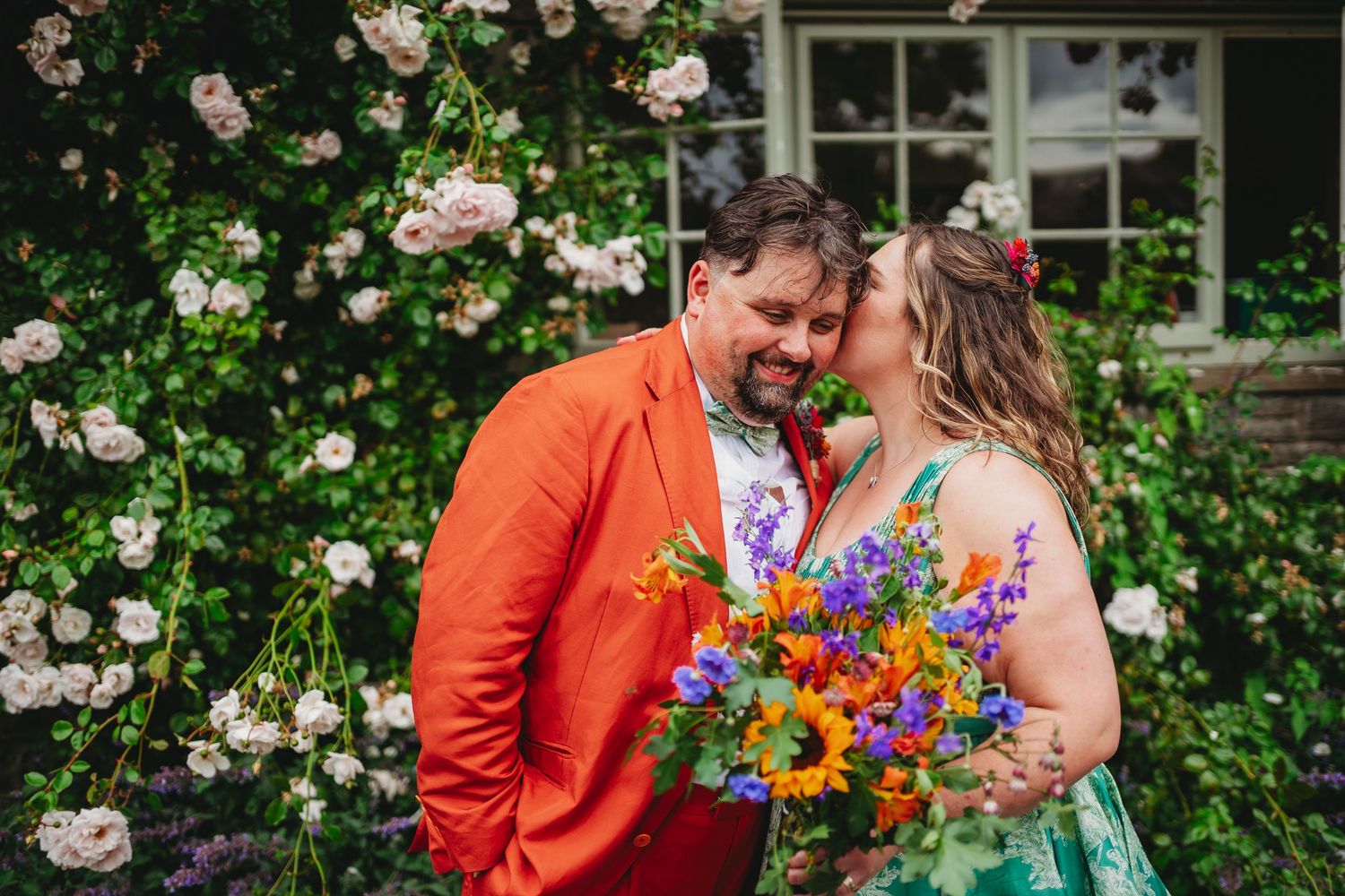 Couple in colorful wedding attire kissing by floral arch, groom in orange suit, bride holding vibrant sunflower bouquet at Glanusk Estate