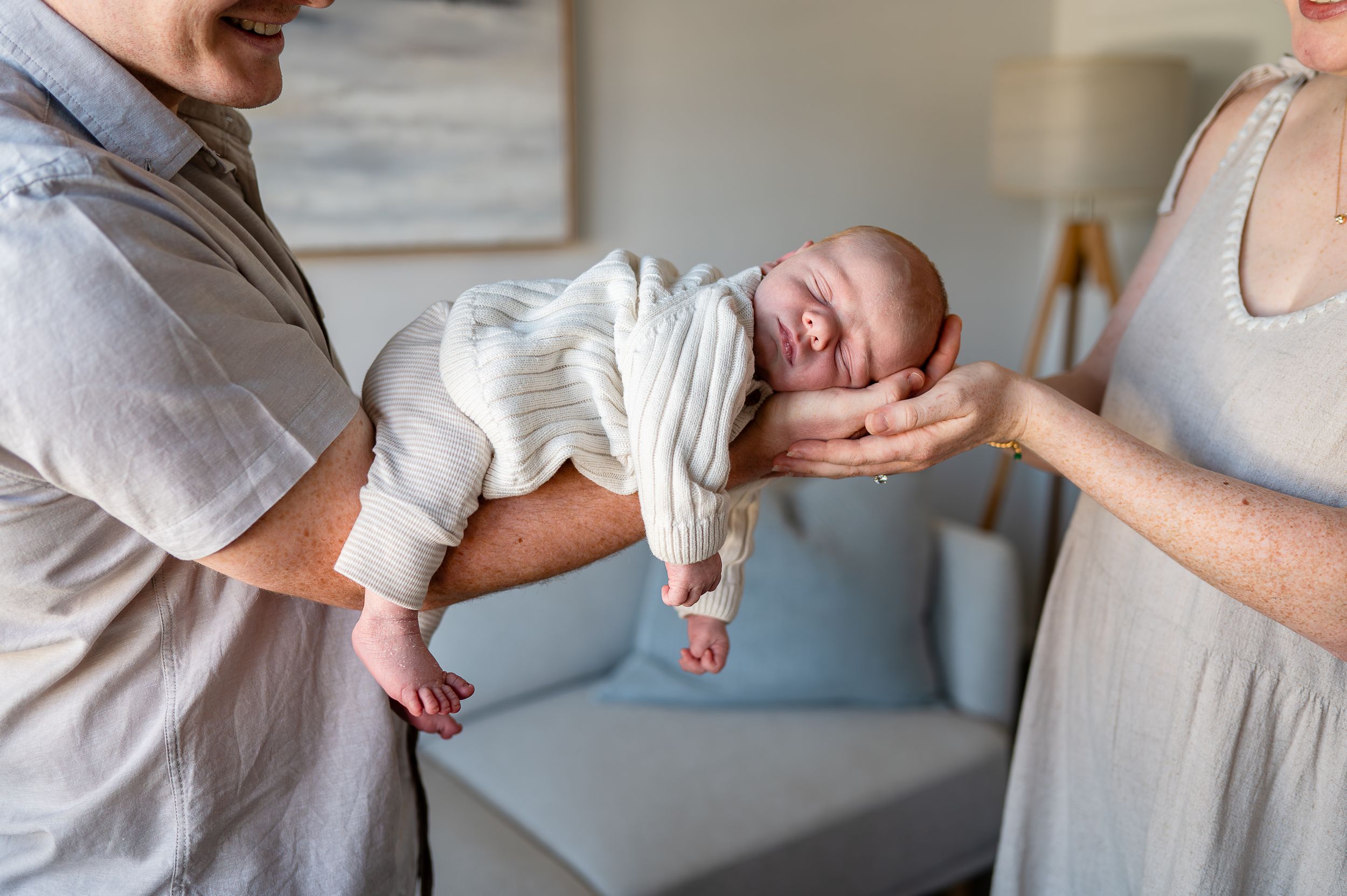 Parents gently holding their swaddled newborn baby together in a bright, cozy living room.