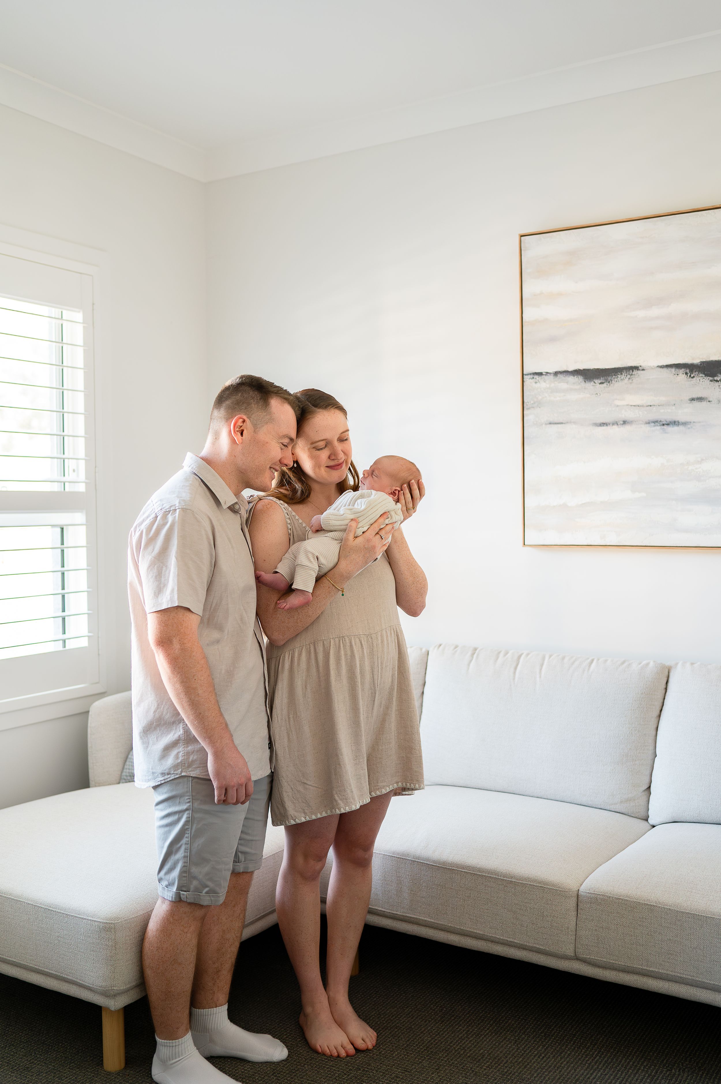 New parents standing in bright living room, lovingly gazing at their newborn baby, with abstract art on wall.