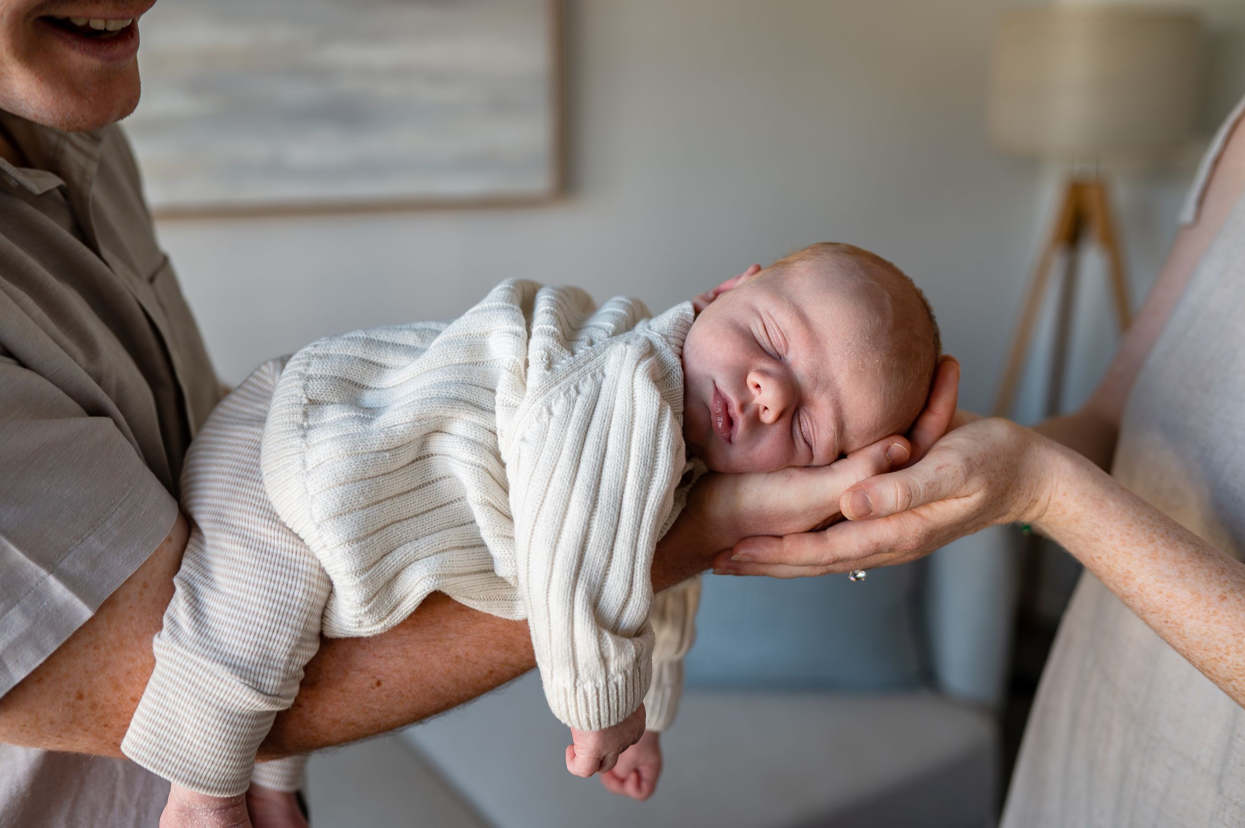 Newborn baby sleeping peacefully in parents hands, wrapped in a cream knit outfit during a lifestyle newborn photo session.