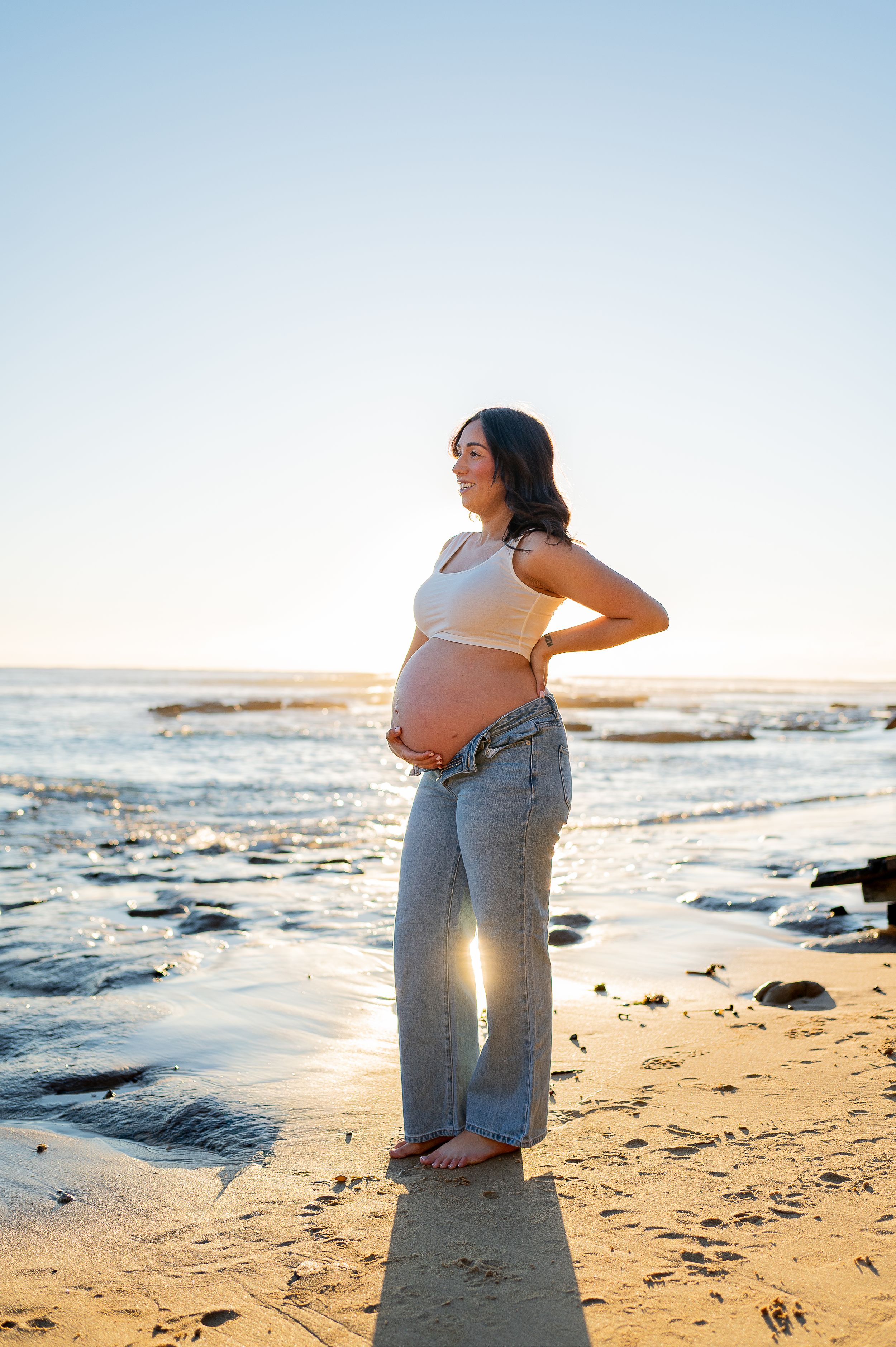 Smiling pregnant woman in crop top and jeans cradling baby bump at golden hour on a sunny beach.