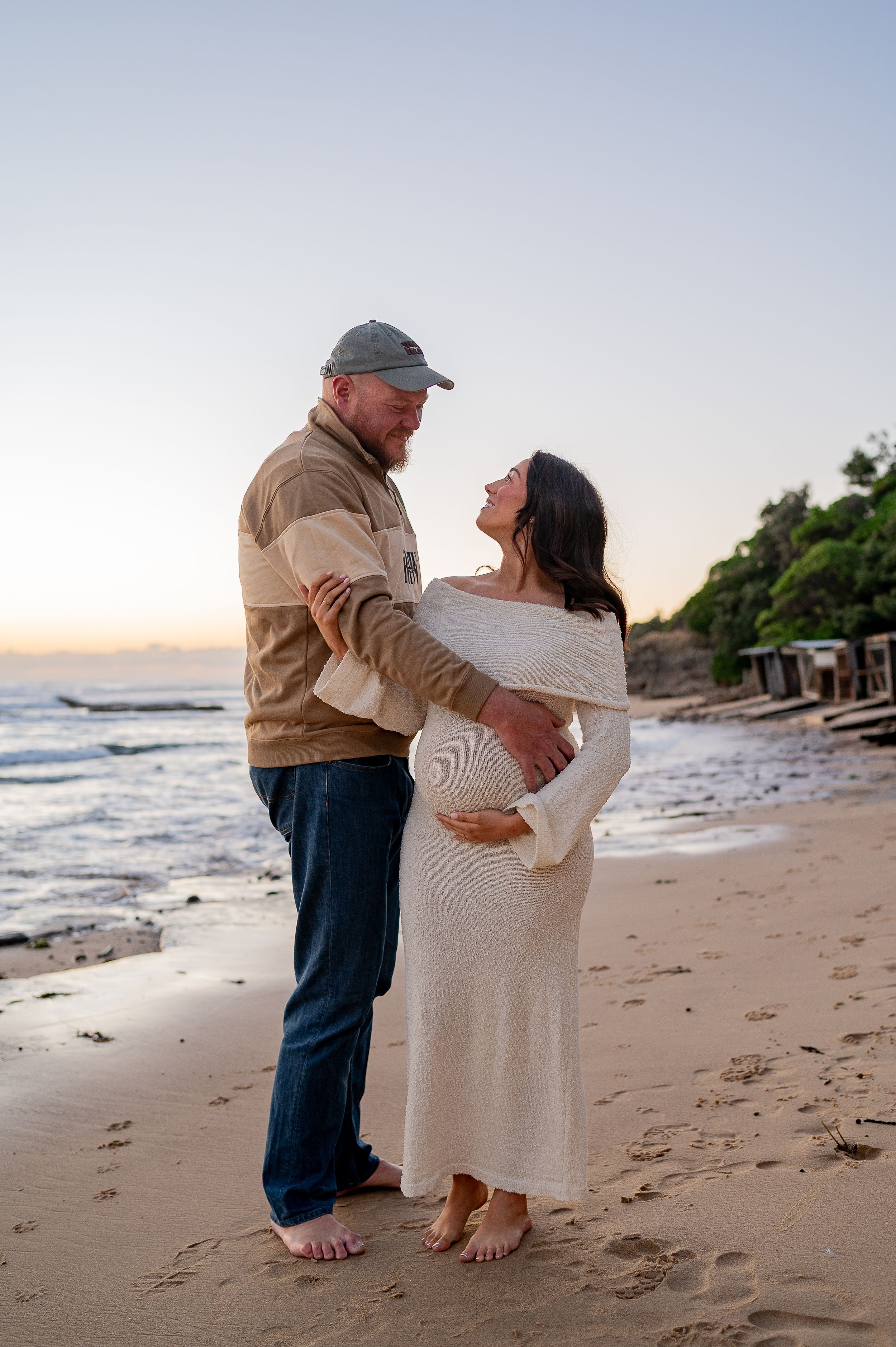 Couple sharing a tender moment on a sandy beach at sunset, woman in white off-shoulder maternity dress cradling her bump.