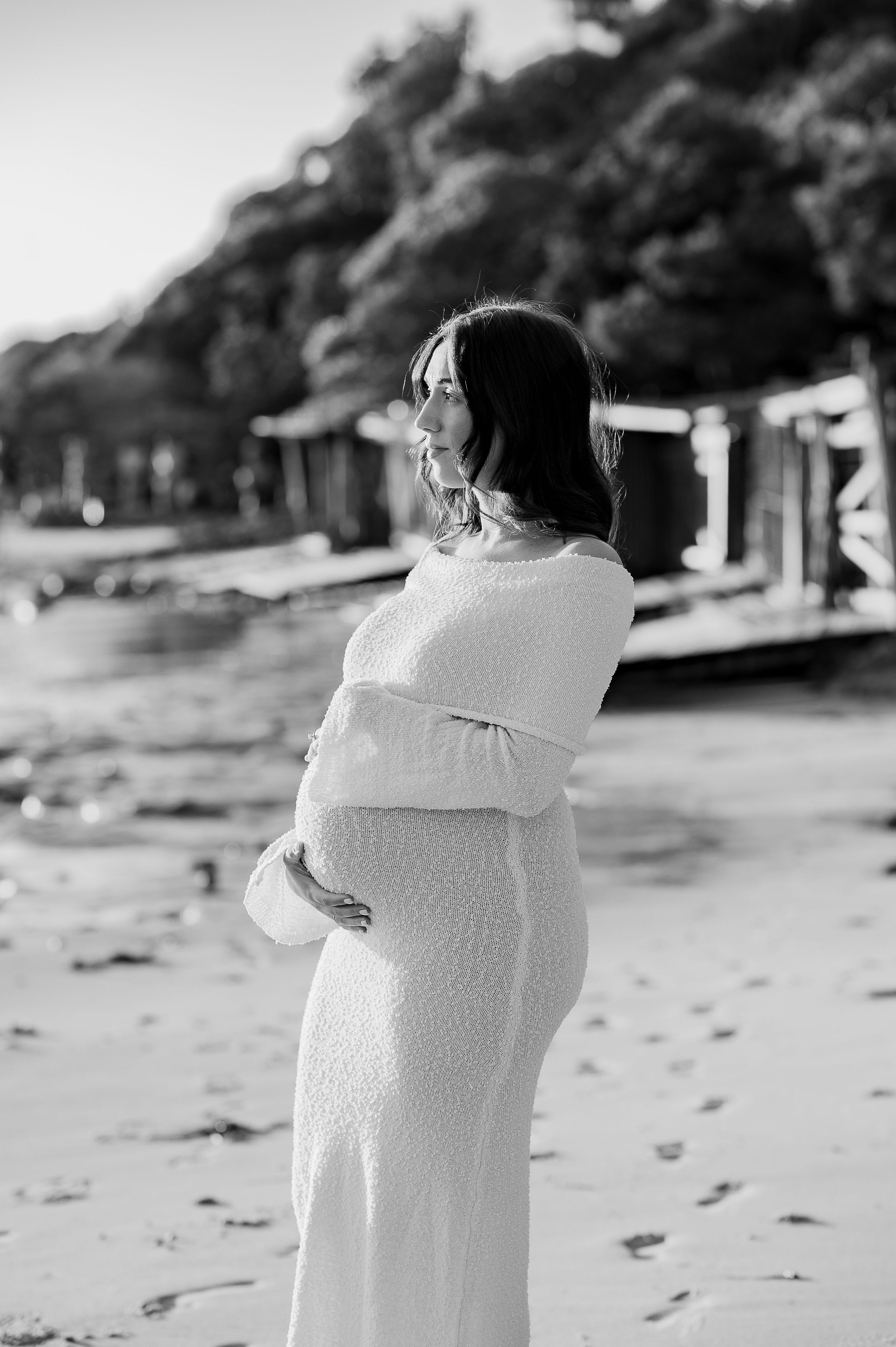 Pregnant woman in white knit dress cradling baby bump on sandy beach in black and white photo.
