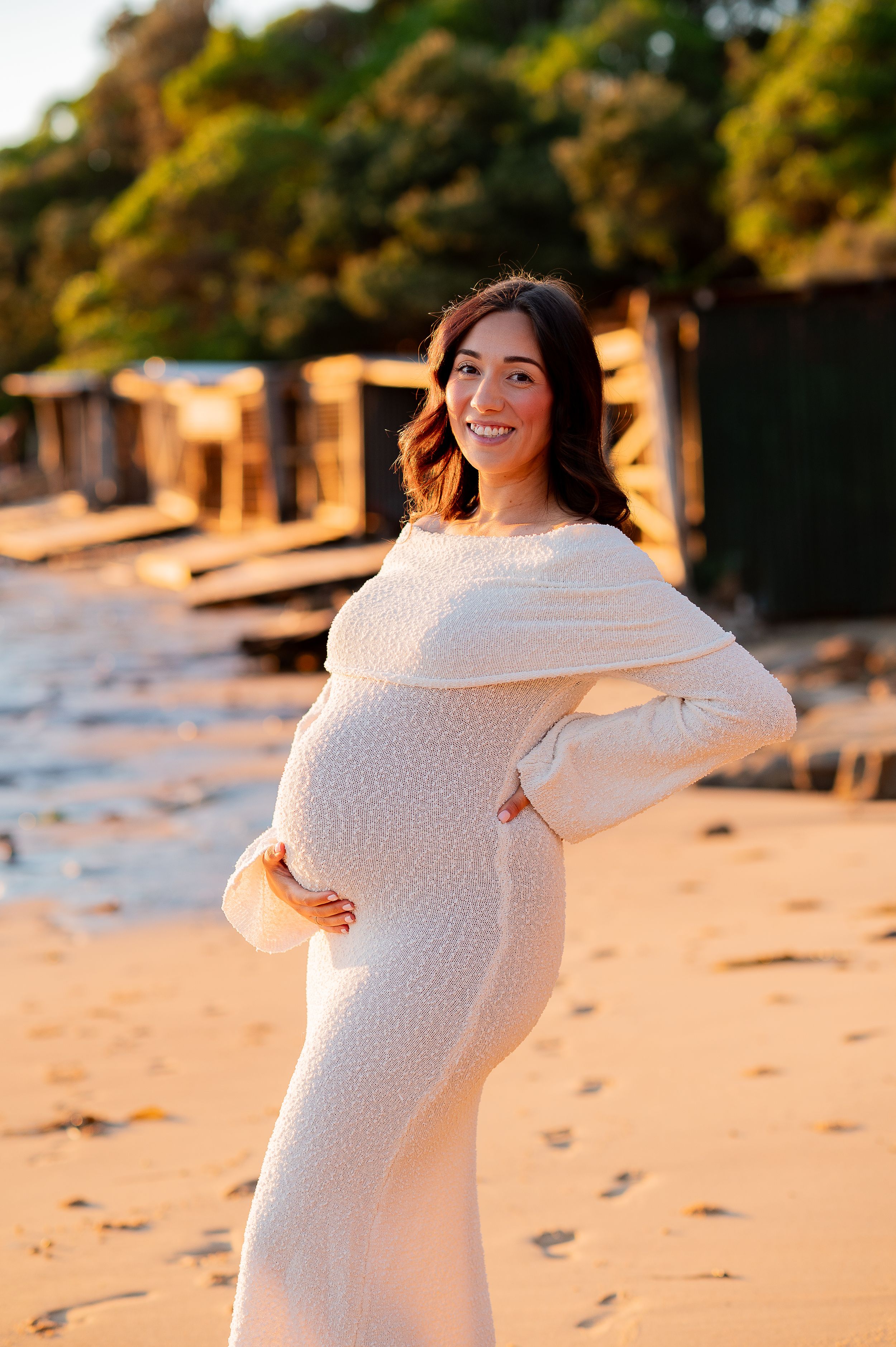 Smiling pregnant woman in white knit dress cradling baby bump on sandy beach at golden hour sunset.
