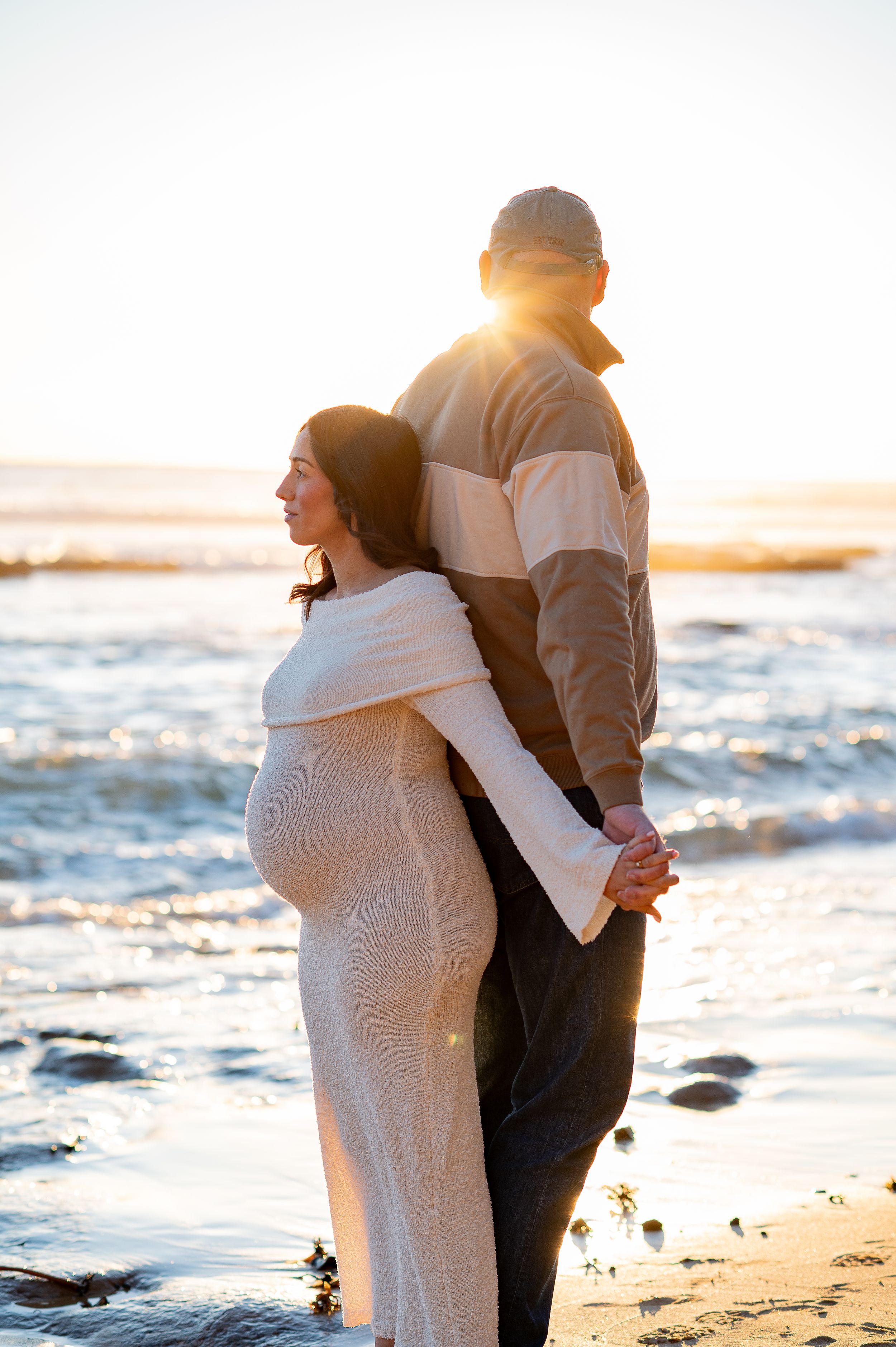 Pregnant couple holding hands at sunset on the beach, woman in white knit dress, man in striped jacket.