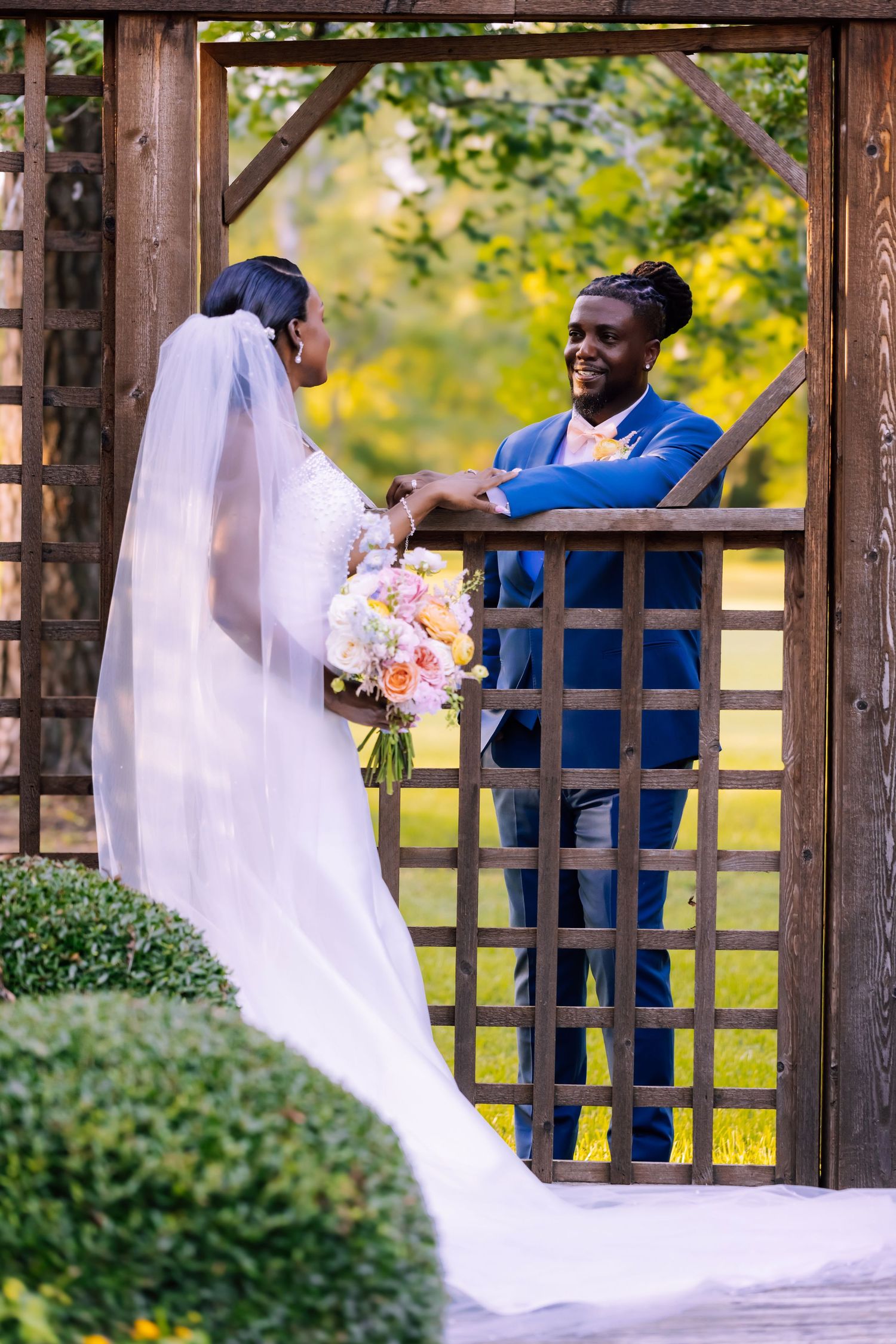 Bride in a white strapless gown holding a colorful bouquet stands in a sunlit park as the groom in a blue suit leans against a tree.