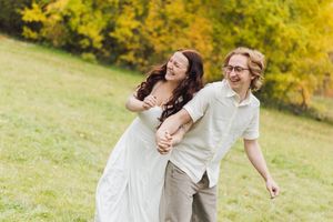 A laughing couple in light summer clothes holds hands and plays together on a sunny grassy hillside with golden autumn trees behind.