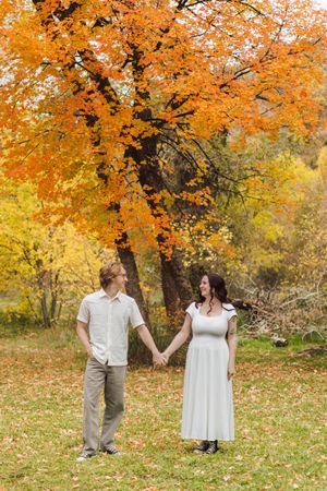 Couple holding hands and smiling at each other beneath a vibrant orange maple tree in an autumn park with fallen leaves.