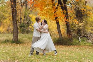 Couple in a white dress and light shirt embrace on a grassy lawn surrounded by vibrant orange and yellow autumn trees.