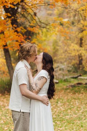 Couple kissing among golden autumn trees, the woman in a white dress with arm tattoos and the man in a white shirt.