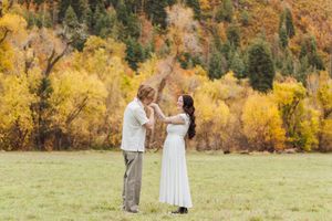 Couple in a white dress and light shirt share a tender moment in a grassy meadow framed by golden autumn trees and mountain hillside.