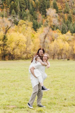 Smiling couple in white outfits and sneakers enjoy a piggyback ride in a grassy meadow with golden autumn trees behind them.