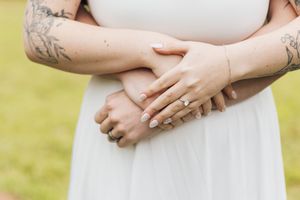Close-up of a tattooed couple's clasped hands resting on a white dress, one wearing a diamond engagement ring, on a blurred green lawn.