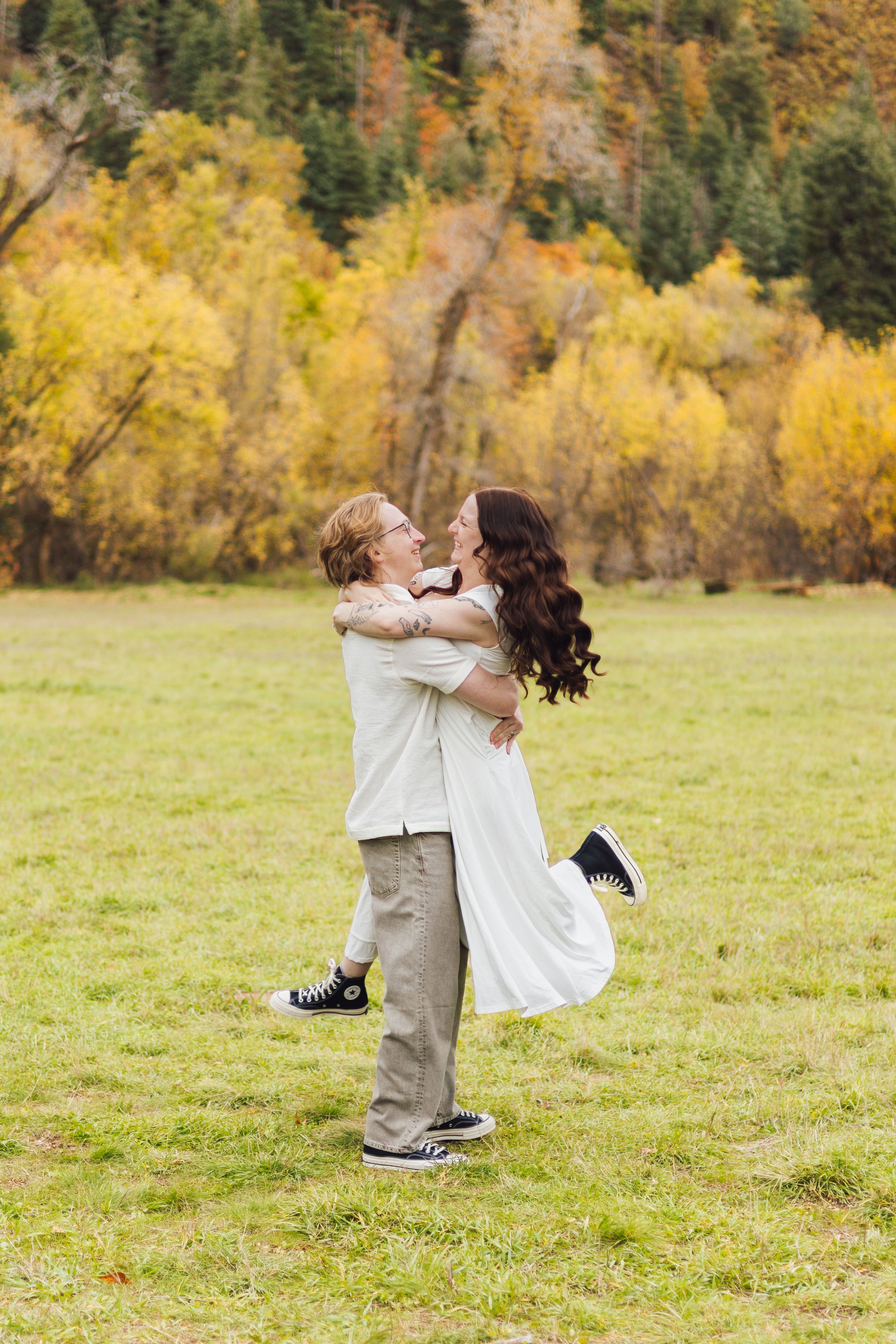 Couple embracing in a grassy meadow as he lifts her off the ground, with golden autumn trees in the background.