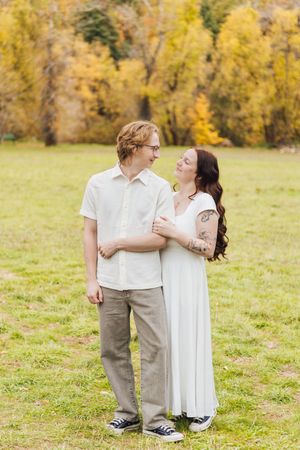 Couple in white outfits embracing and smiling at each other in a grassy field with golden autumn trees in the background.