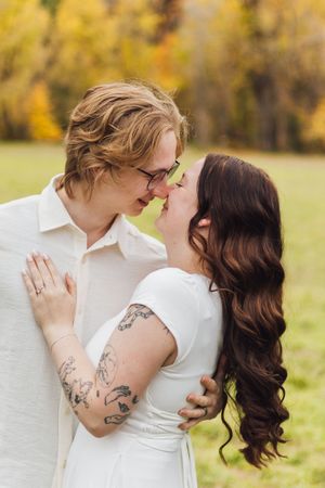 Couple in white outfits touch noses in an affectionate embrace in a grassy field with golden autumn trees behind them.