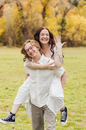 Smiling couple in an autumn park, a woman with tattoos piggybacking on a glasses-wearing man in a white shirt and black sneakers.