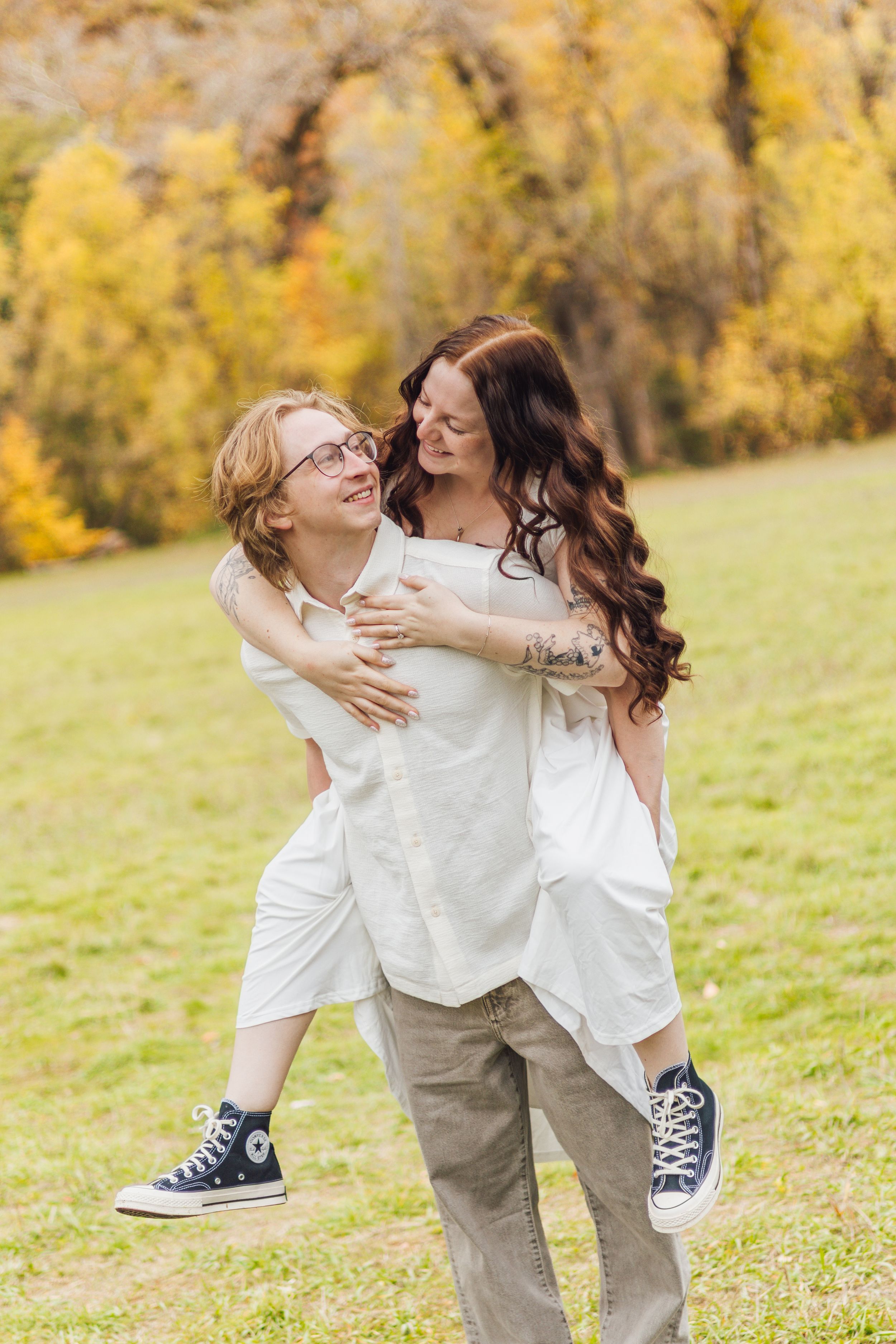 A smiling couple poses outdoors as a woman with long wavy hair rides piggyback on a man in glasses, with autumn trees in the background.