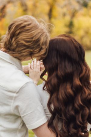 Couple embracing from behind with heads together and hands touching, showing a ring, in a golden autumn park.