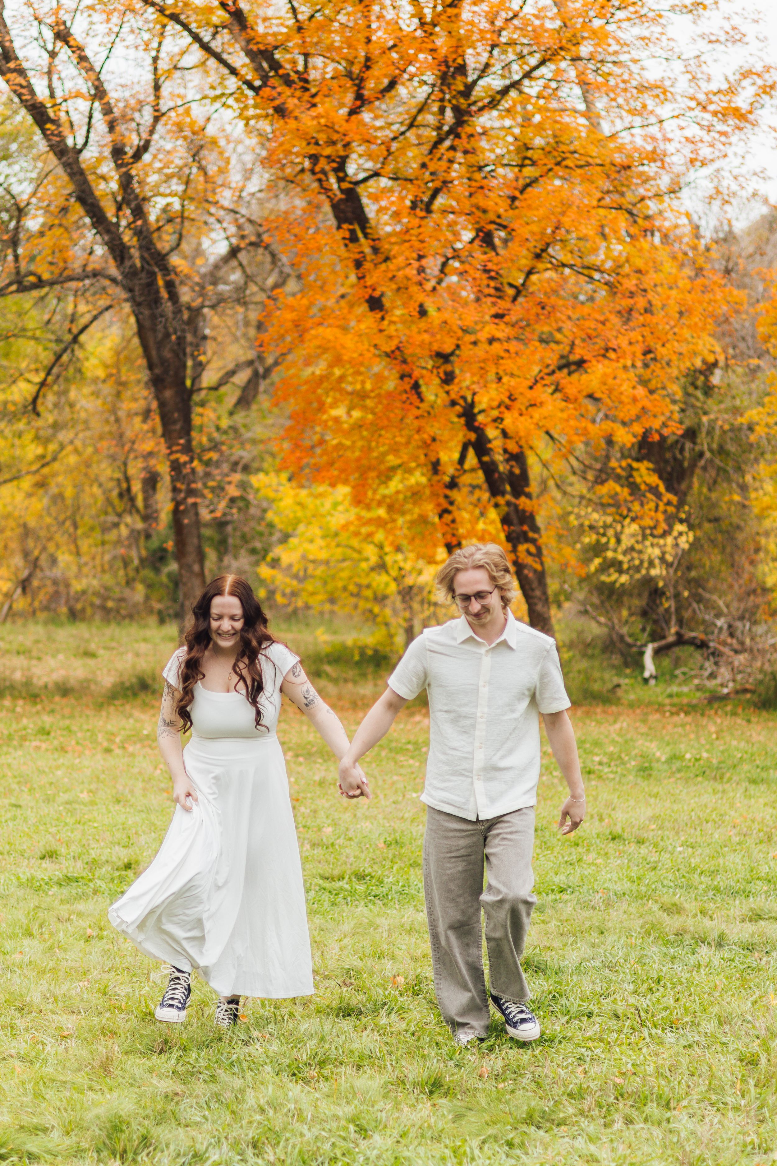 Couple holding hands and walking through a grassy field beneath vibrant orange autumn trees, the woman in a white dress and the man in a but