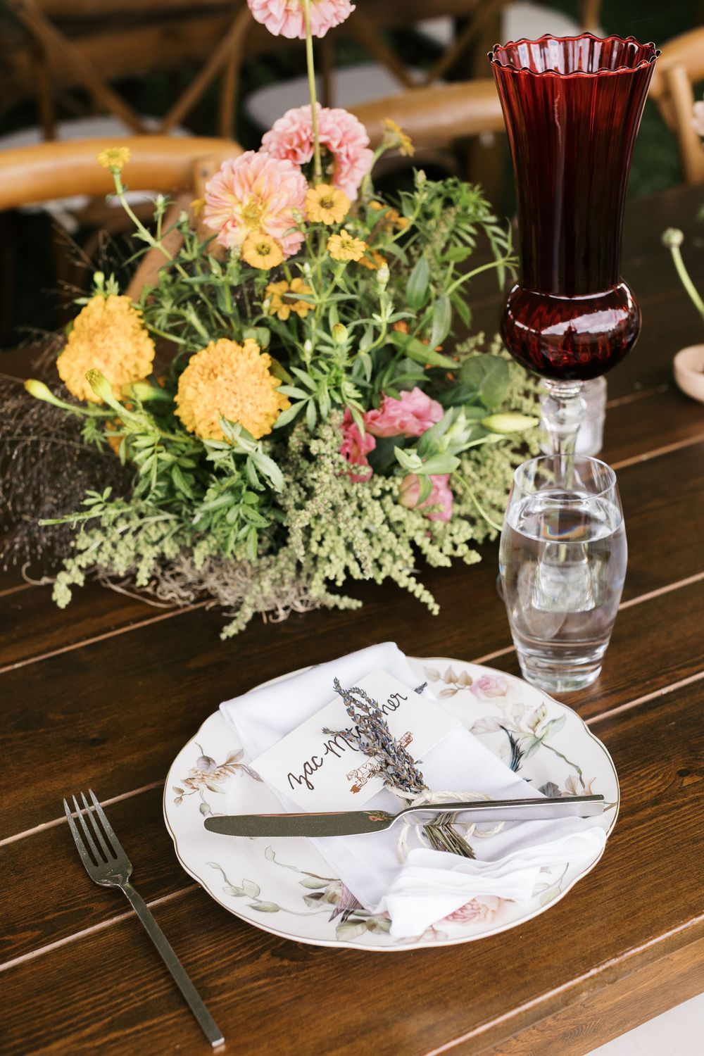 Elegant wedding table setting with floral centerpiece, ruby red goblet, and botanical-print china on dark wood table.