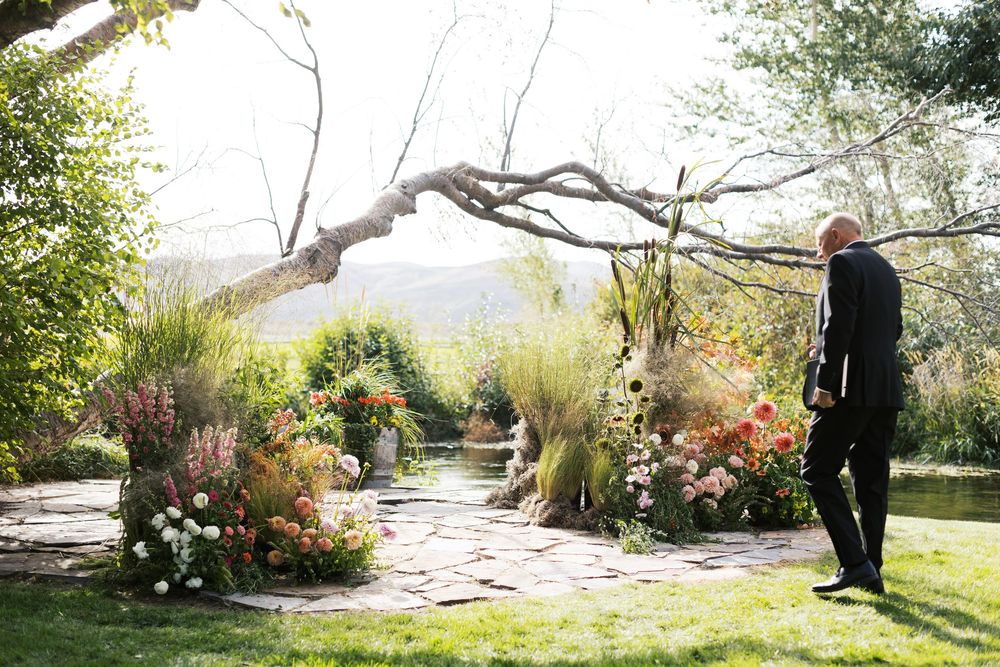 Wedding photographer capturing a floral ceremony arch surrounded by lush pink blooms and ornamental grasses outdoors.
