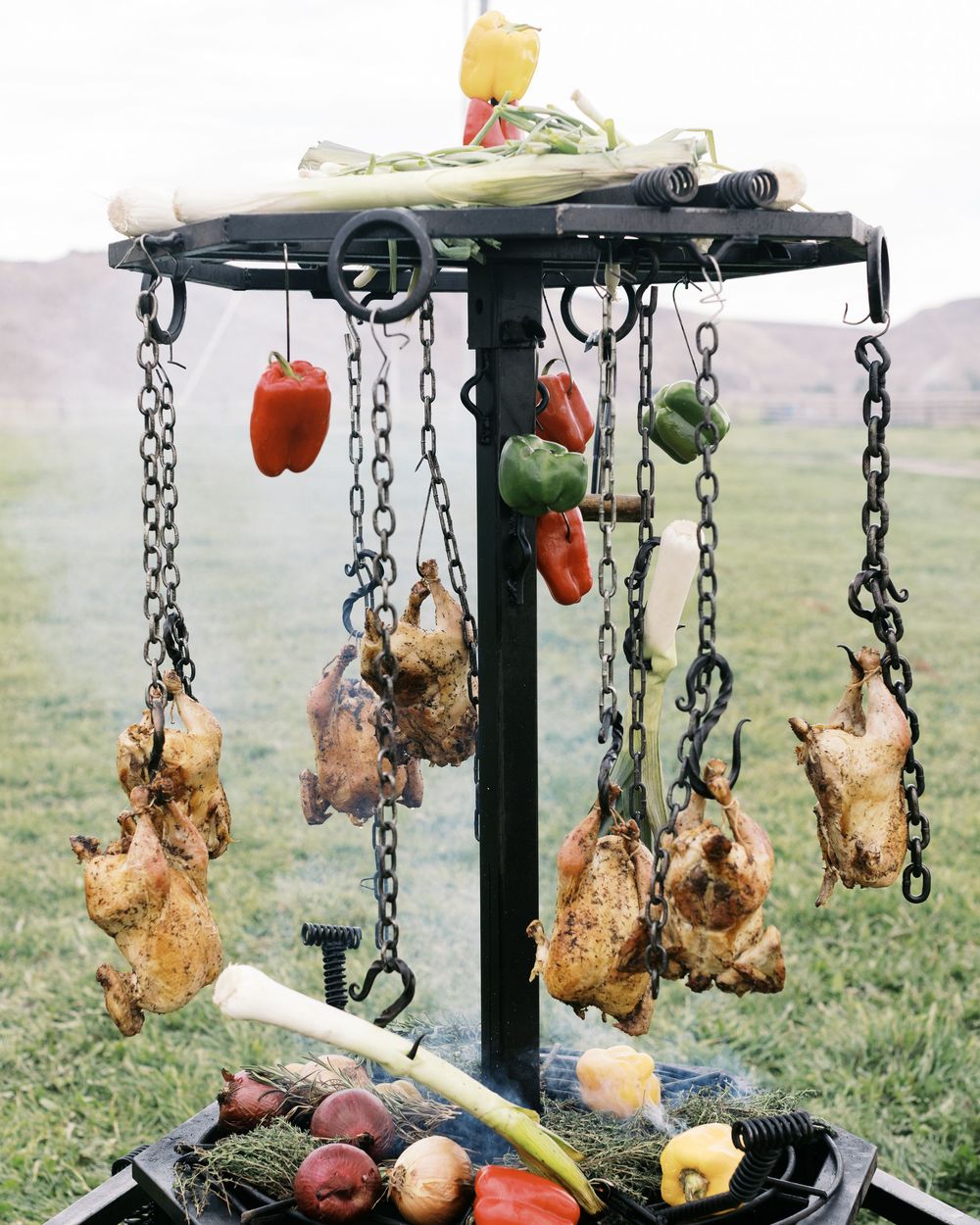 Outdoor rotisserie grill with whole chickens and colorful peppers hanging from chains over an open fire.