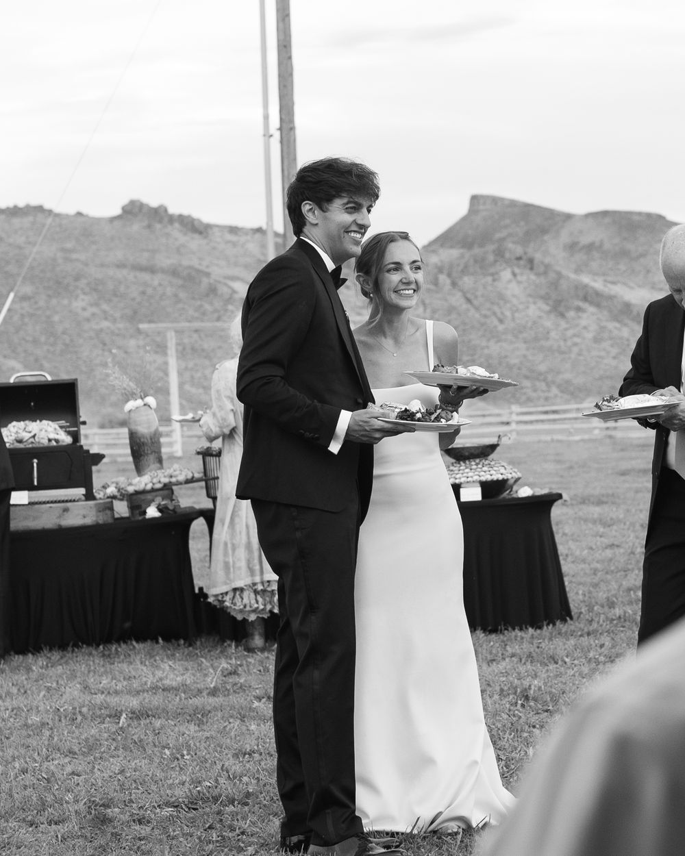 Happy bride and groom smiling while holding plates at their outdoor wedding reception buffet in black and white.