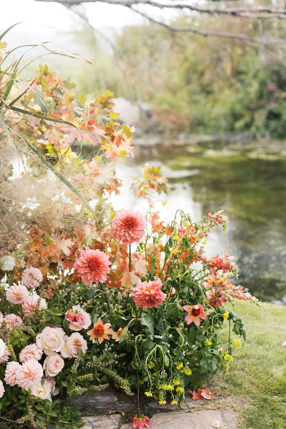 A romantic floral arrangement of pink dahlias and roses sits beside a tranquil reflective pond.