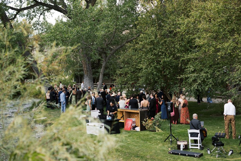 Outdoor wedding cocktail hour with guests mingling under large trees beside a scenic river setting.