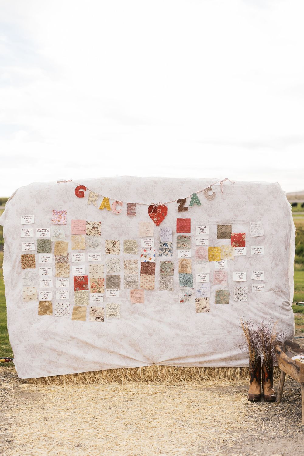 Rustic wedding seating chart displayed on a white quilt draped over hay bales with colorful fabric squares and bunting.