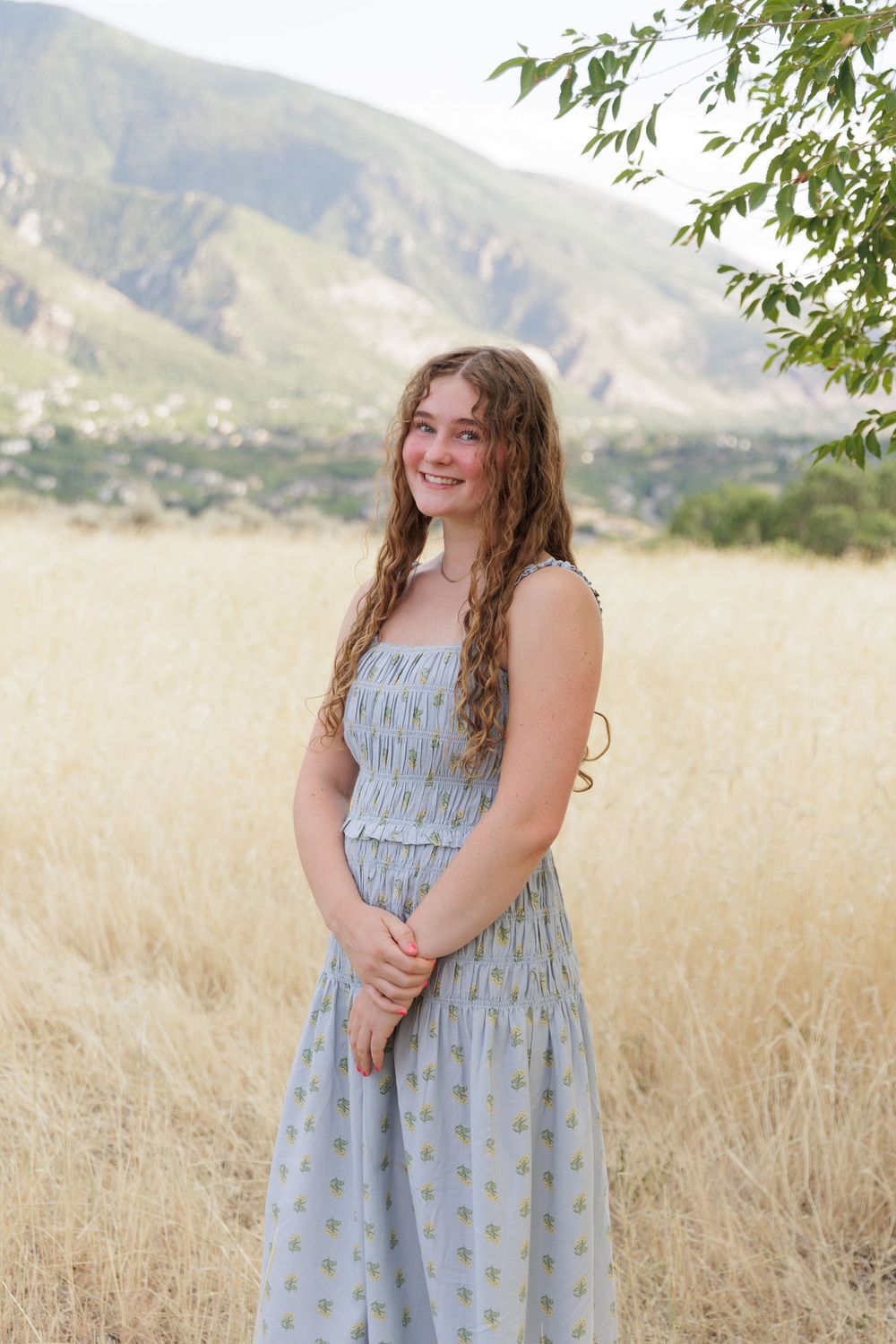 Young woman in blue floral sundress smiling in golden field with mountain backdrop.