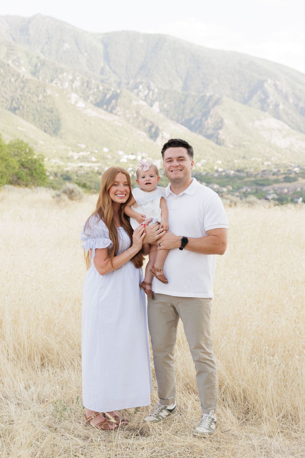 Happy young family with baby poses in sunny golden field with mountain views.