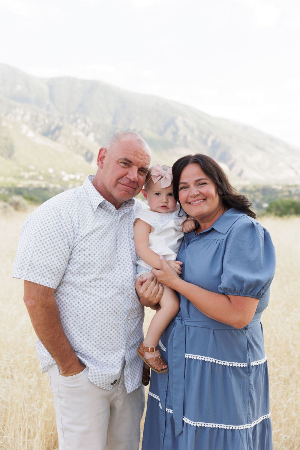 Grandparents smiling and holding adorable baby in a golden field with mountains.