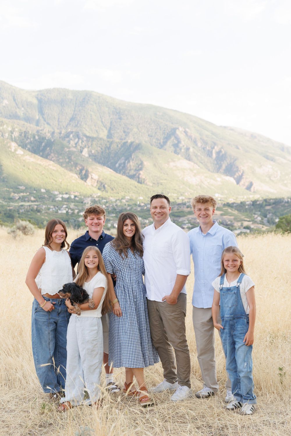 Large family of seven with dog poses in golden field with mountain backdrop.