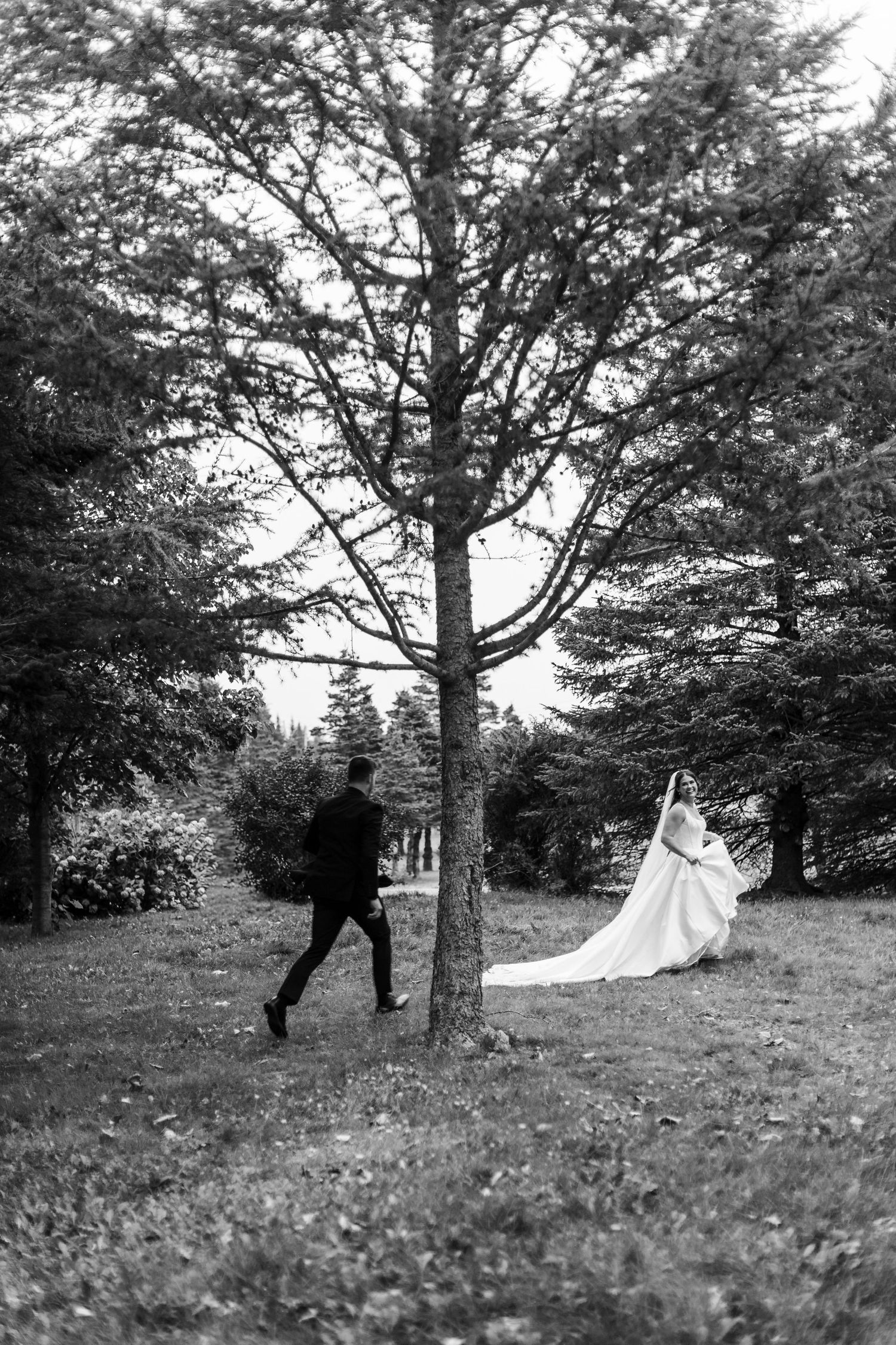 Black and white wedding photo of a groom walking toward a bride in a long gown with a flowing train among tall trees on a grassy lawn.