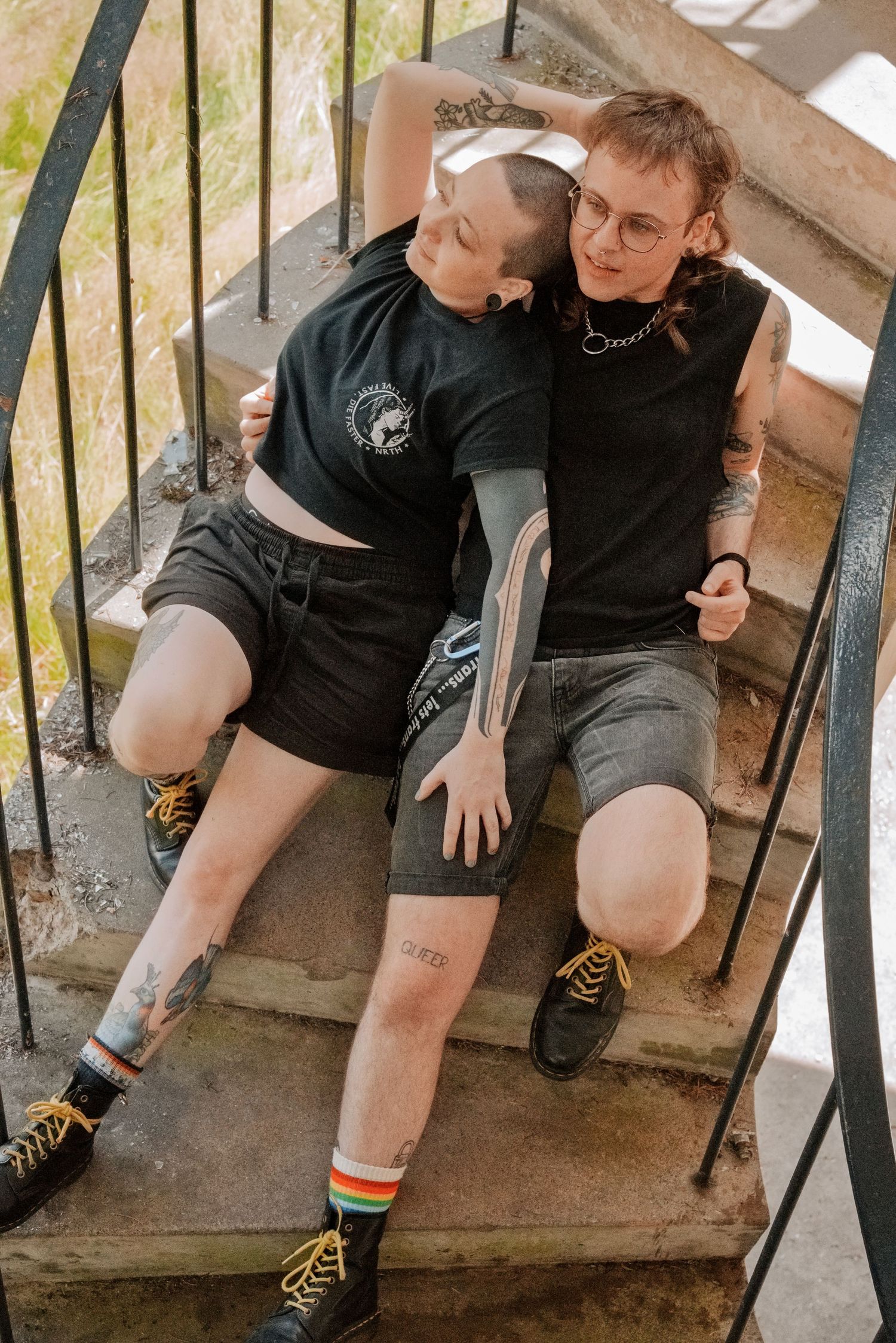 Two tattooed people in black outfits sitting together on outdoor stairs, wearing shorts and boots.